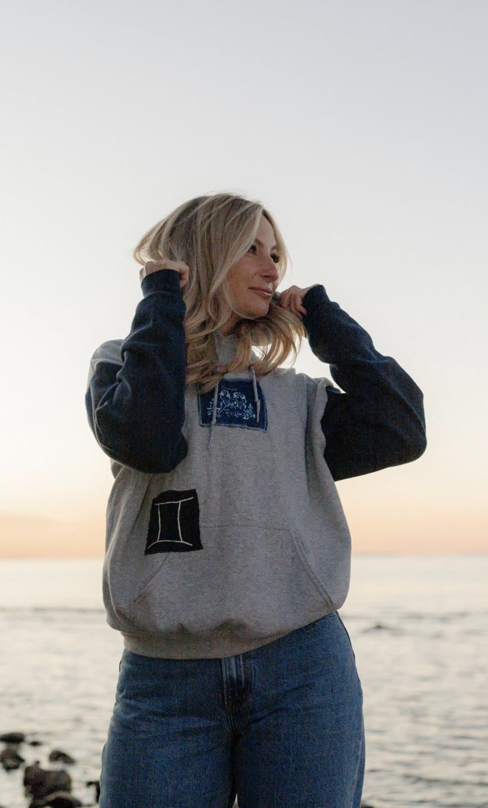 A grey hoodie with navy sleeves and denim patchwork, on the beach at sunset